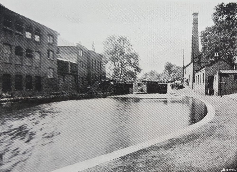 1950s black and white photo of canal, large chimney on right of image. Factories on left of image.