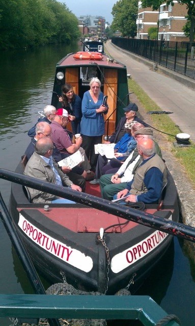 11 people sit on the front of a canal boat named opportunity. Group is turned to listed to woman (Carolyn Clark).