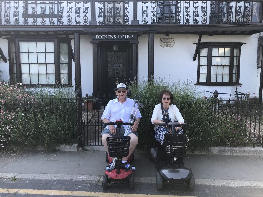 Dennis and Ellen in motorised wheelchairs in front of a black and white building. Dickens House written above the door.