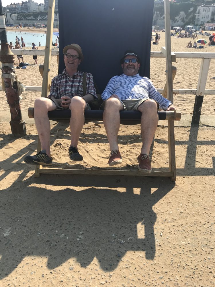 John and Paul sit side by side in an oversized deck chair. Wearing hats and shorts.